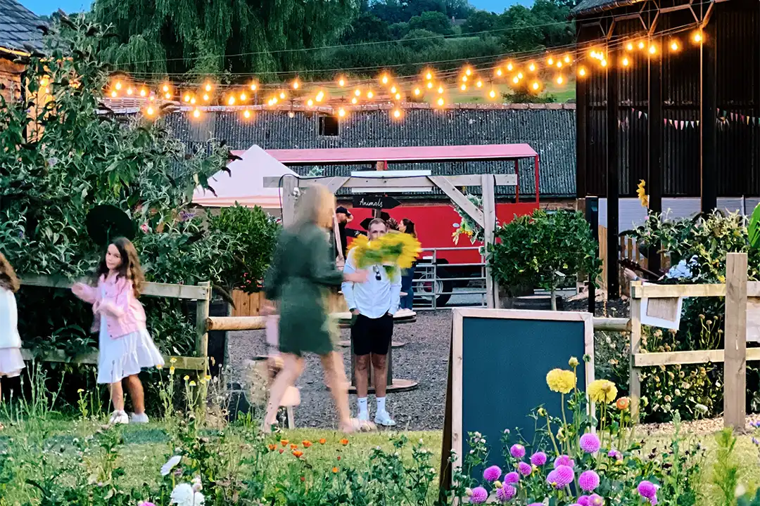 A crowd moves amongst the farm courtyard at dusk, lit by festoon lights
