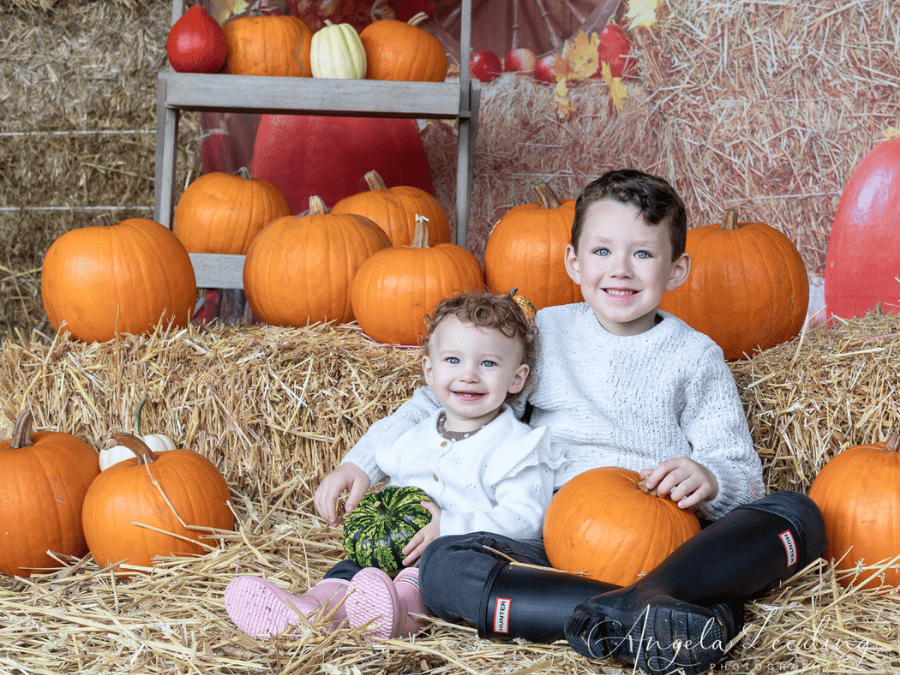 Two smiling young children sitting on straw among pumpkins during an autumn barn photoshoot.